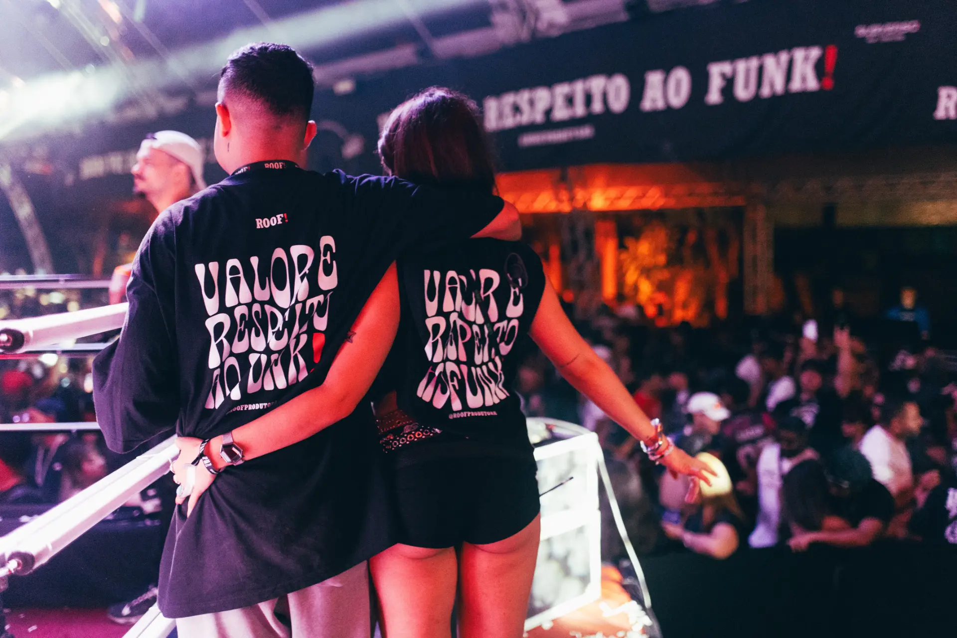Casal na festa ROOF! vestindo as camisetas oficiais, com faixa 'RESPEITO AO FUNK!' ao fundo